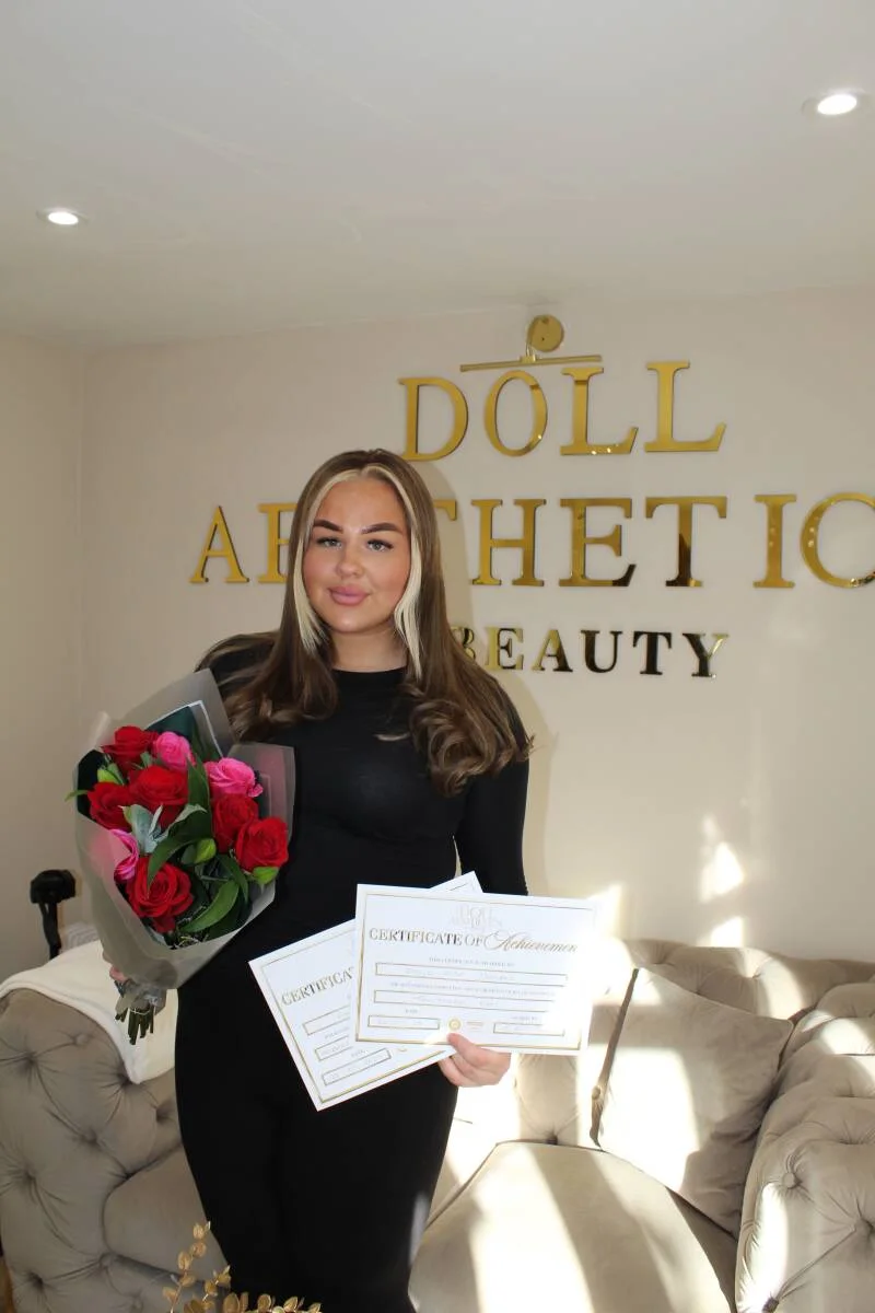 Doll Academy graduate holding her certificate and flowers by the gold clinic signage